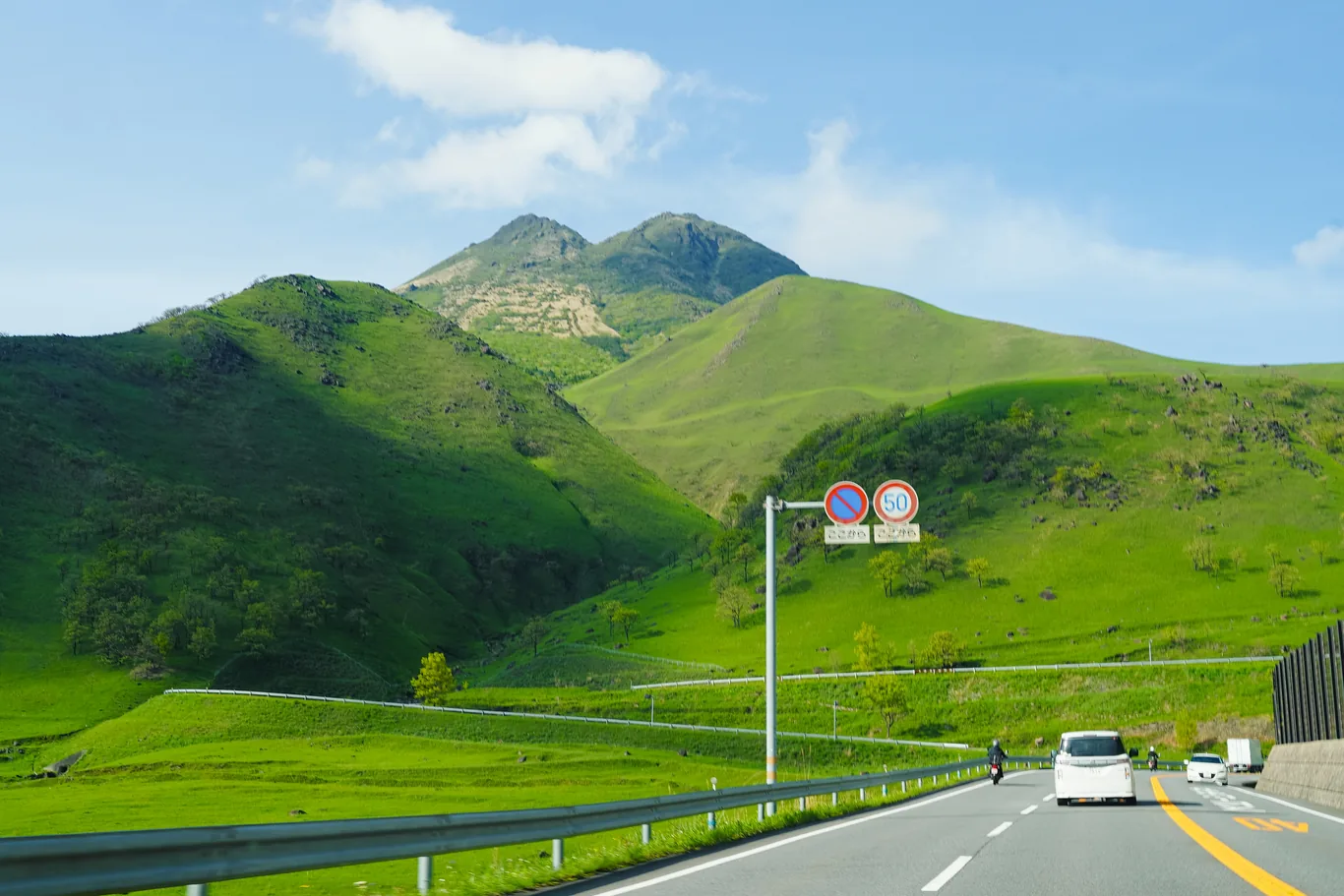 Zweispurige Straße vor grünen Hügeln, dahinter der Doppelgipfel des Yufu-dake, rechts zwei runde Verkehrsschilder