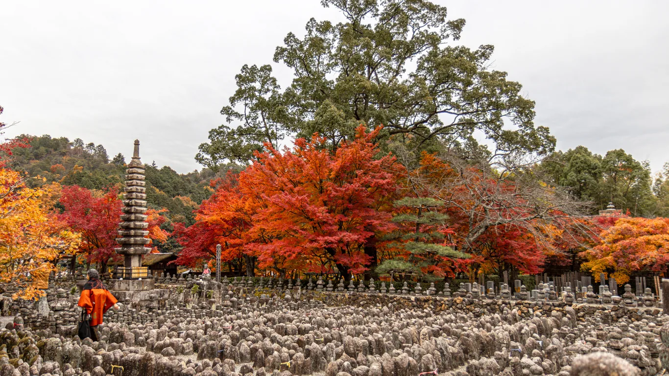 Feld aus Tausenden kleinen Steinfiguren vor einer Steinpagode, dahinter rotes Herbstlaub