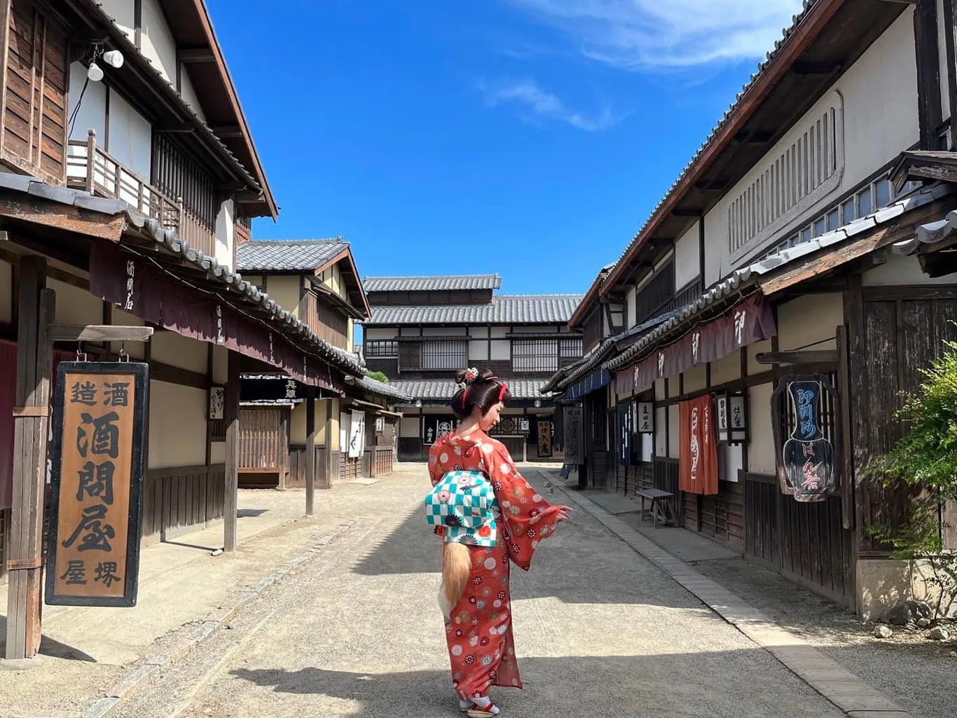 Frau in rotem Kimono mit türkisem Obi und Fuchsschwanz von hinten auf einer leeren Kulissenstraße unter blauem Himmel