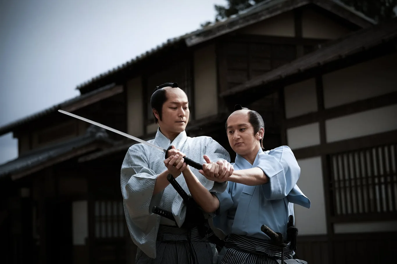 Zwei Schauspieler in Kimono und Hakama mit Samurai-Frisur ringen um ein Schwert, dahinter dunkle Holzhäuser im Edo-Stil