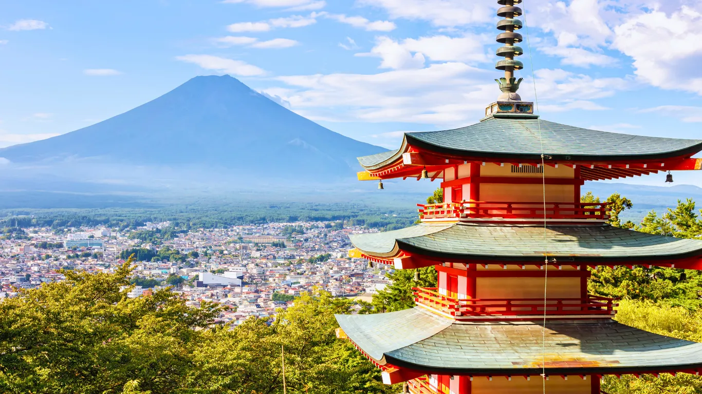 Eine rote Pagode im Vordergrund, dahinter eine Stadt und der Fuji im Sommer ganz ohne Schnee