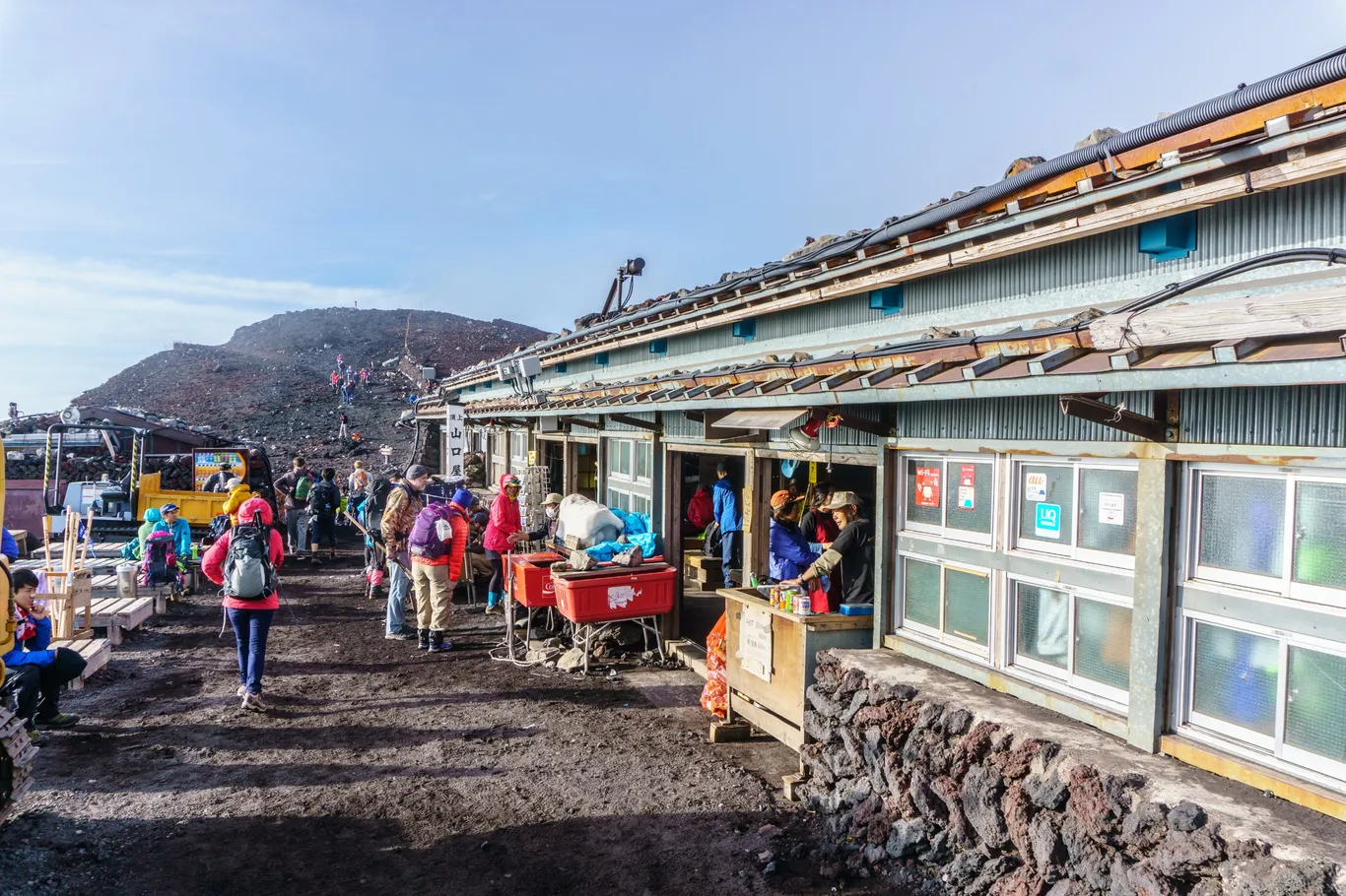 Eine flache Berghütte aus Holz und Wellblech am Gipfel, davor Bergsteiger in Funktionsjacken, im Hintergrund ein Weg auf einen Kraterhügel