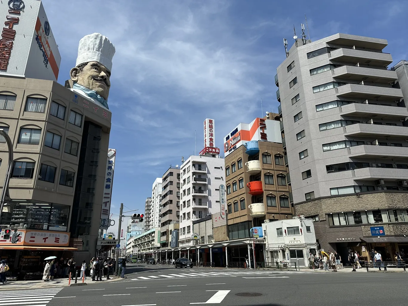 Straßenkreuzung in Kappabashi: links ein Gebäude mit einem riesigen Kochkopf mit weißer Mütze auf dem Dach, rechts eine Fassade mit großen bunten Kaffeetassen