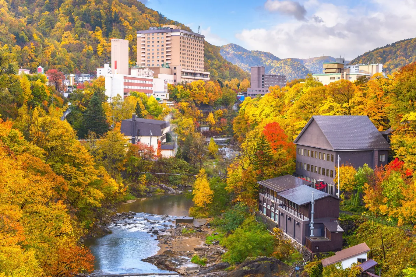 Flusstal mit gelb und orange gefärbten Bäumen, dazwischen Hotels und Häuser des Onsen-Orts Jōzankei, im Hintergrund bewaldete Berge