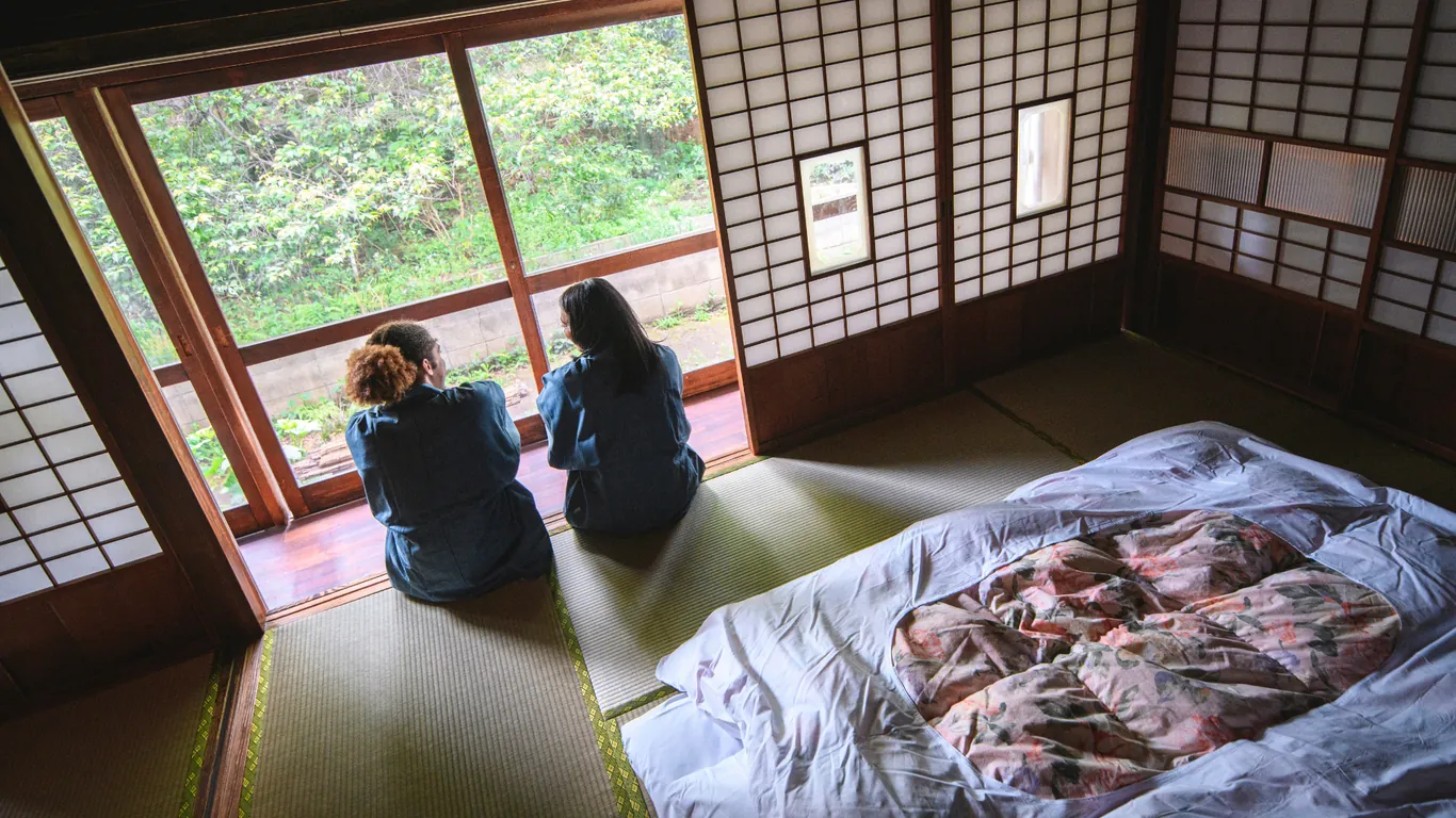 Zwei Personen sitzen von hinten gesehen am offenen Fenster eines Tatami-Zimmers, daneben ein ausgebreiteter Futon