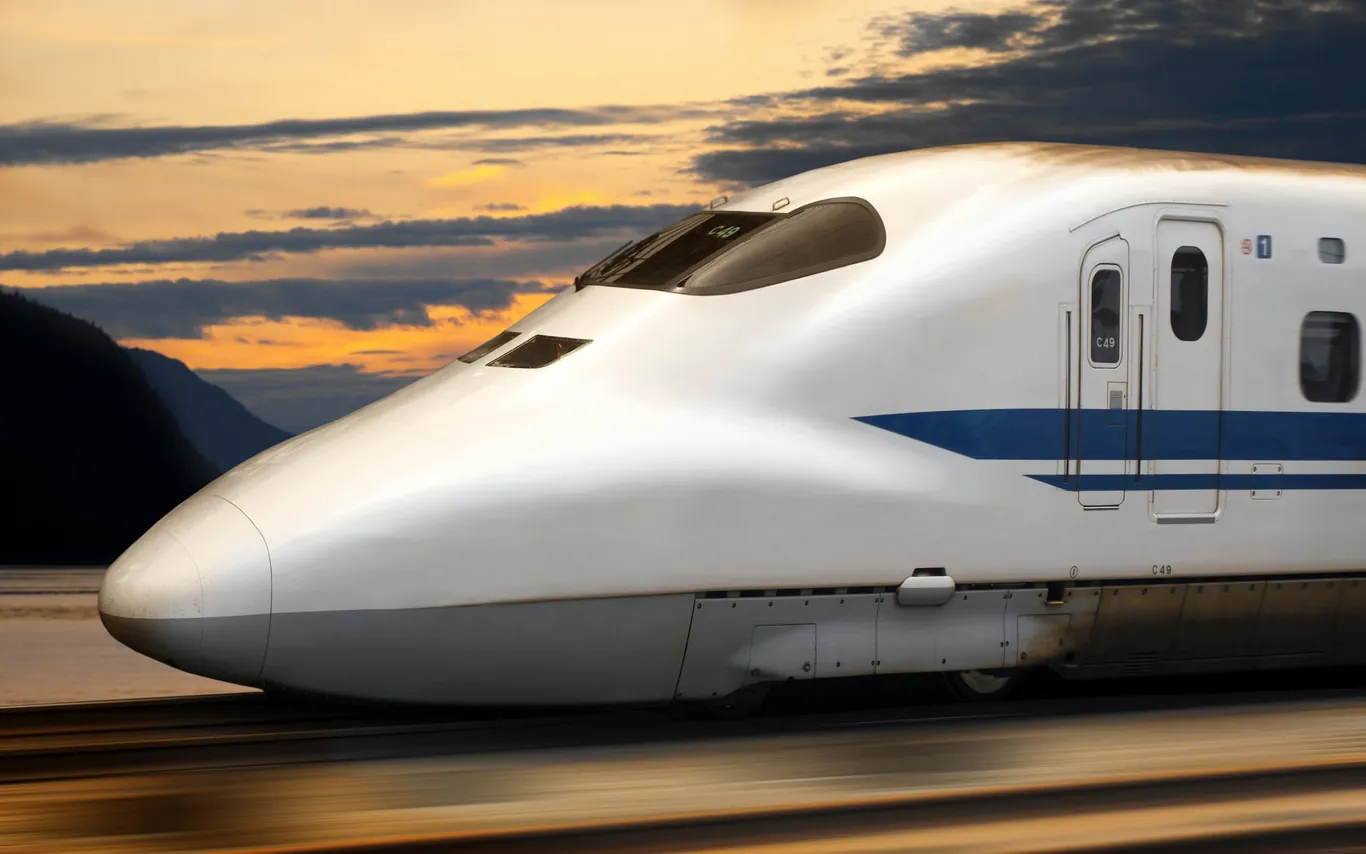 Weißer Shinkansen mit blauen Streifen in der Abenddämmerung, dahinter orange leuchtende Wolken und dunkle Berge