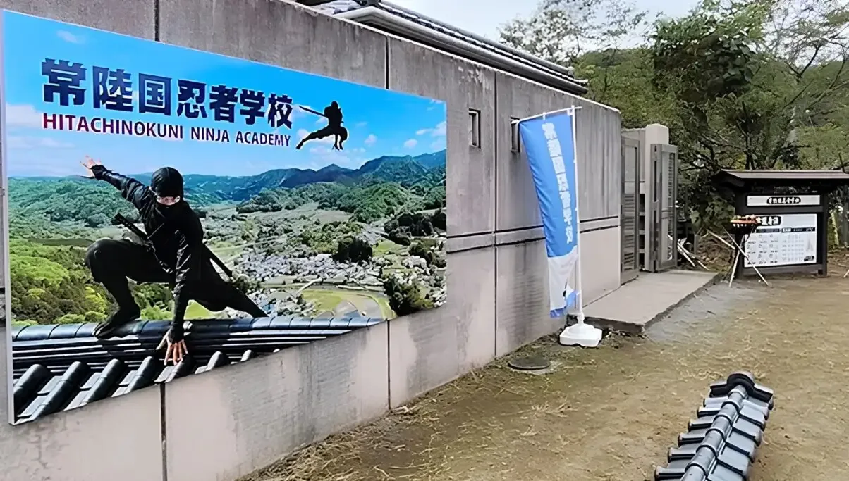 Großes Schild an einer Mauer mit der Aufschrift Hitachinokuni Ninja Academy und einem Ninja auf einem Ziegeldach, daneben eine blaue Fahne