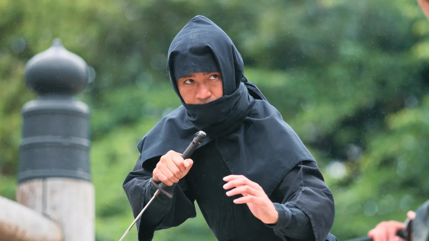 Ein Mann im schwarzen Ninja-Gewand mit Kapuze und Gesichtstuch hält ein Schwert und blickt zur Seite, im Hintergrund grüne Bäume