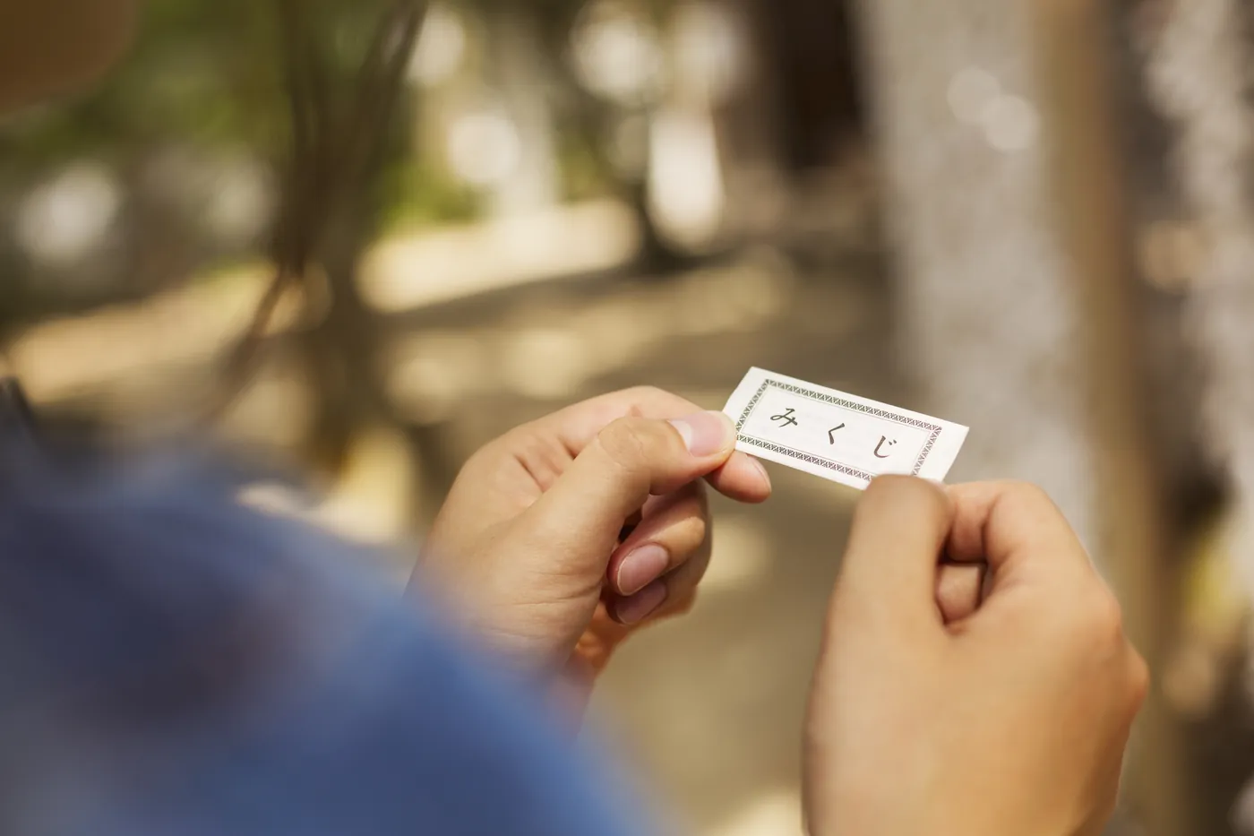 Zwei Hände falten einen schmalen weißen Omikuji-Zettel auseinander, im Hintergrund ein Schreingelände