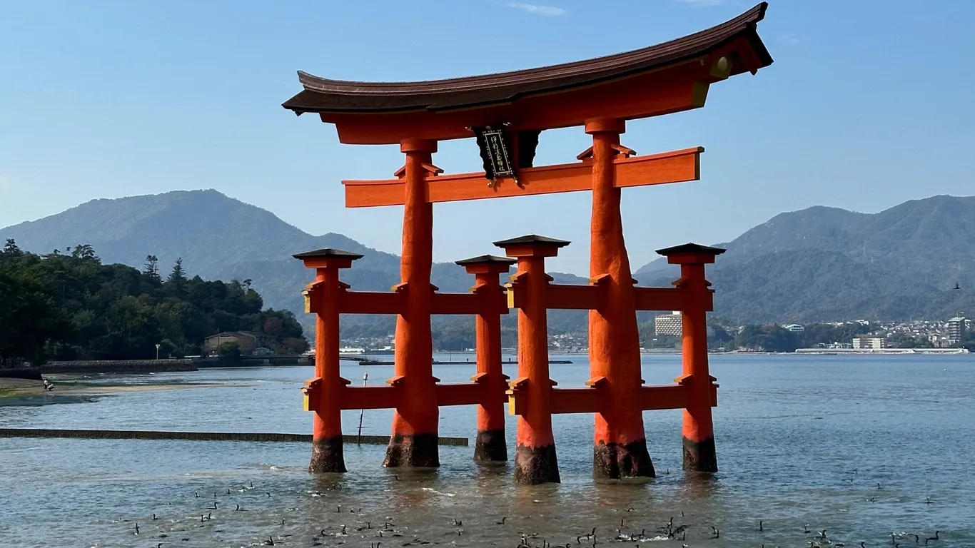 Das große rote Torii von Miyajima im Wasser, dahinter bewaldete Berge und blauer Himmel