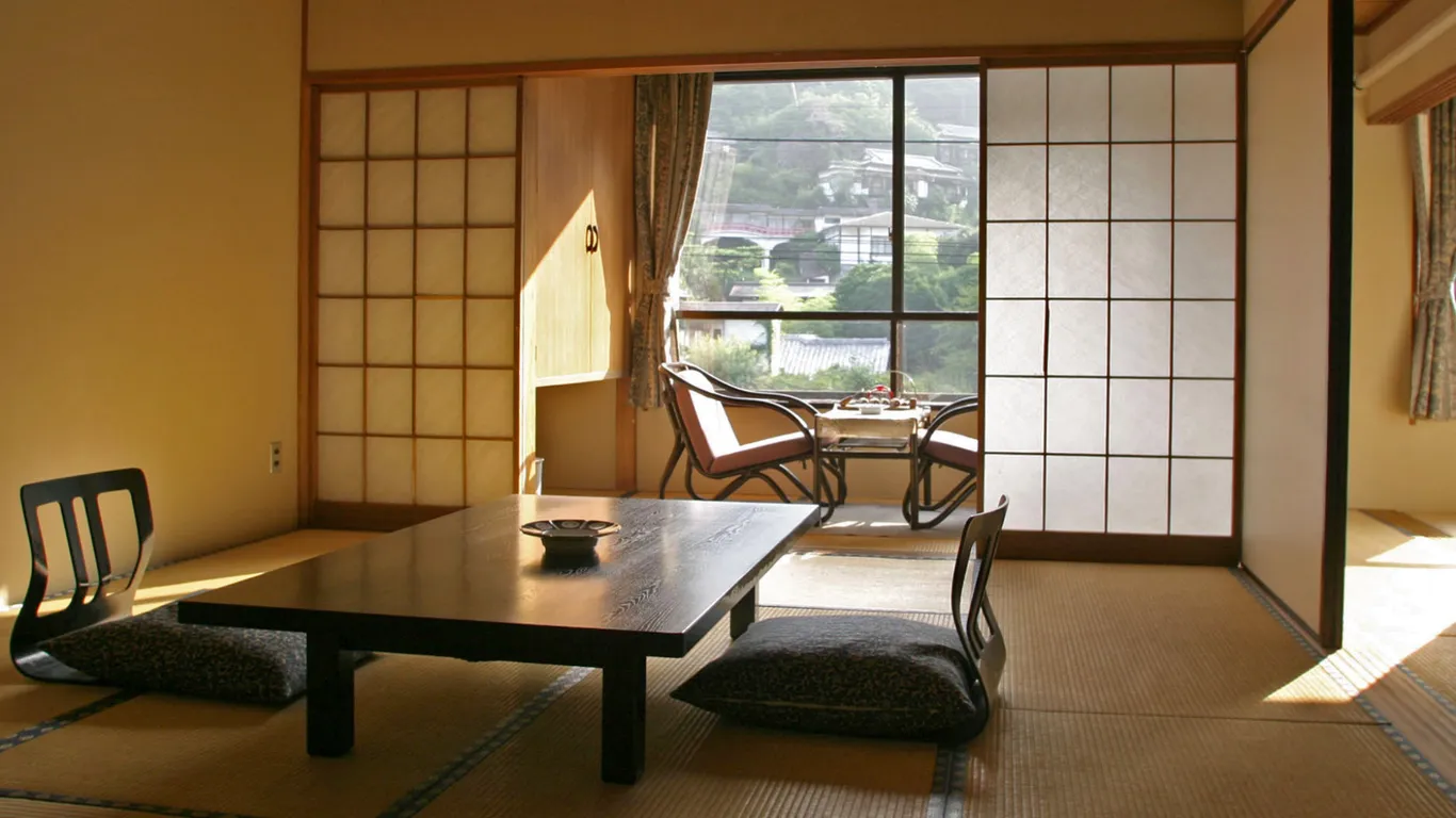 Ryokan-Zimmer mit Tatami, niedrigem Holztisch, zwei Sitzstühlen und Shōji-Schiebetüren, durch das Fenster fällt Sonnenlicht