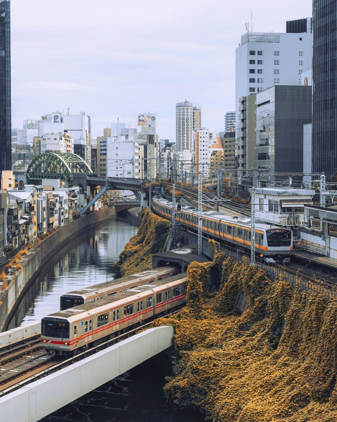 Blick auf den Kanda-Fluss zwischen Häusern, unten ein roter U-Bahn-Zug auf einer Brücke, rechts ein Zug mit orangem Streifen im Bahnhof, hinten eine grüne Bogenbrücke