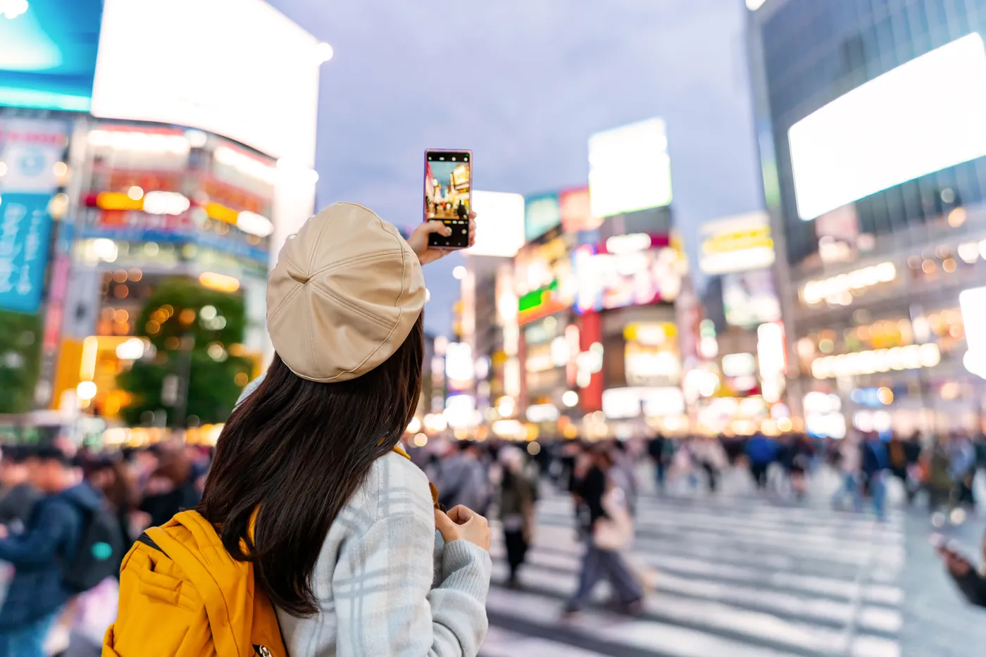 Eine Frau mit heller Kappe fotografiert in der Dämmerung die Leuchtreklamen an der Kreuzung von Shibuya, davor die Menschenmenge auf dem Zebrastreifen