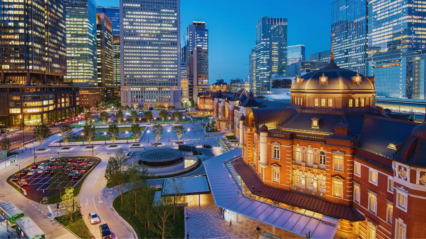 Der beleuchtete Backsteinbau der Tokyo Station in der Abenddämmerung, dahinter Hochhäuser von Marunouchi mit erleuchteten Fenstern