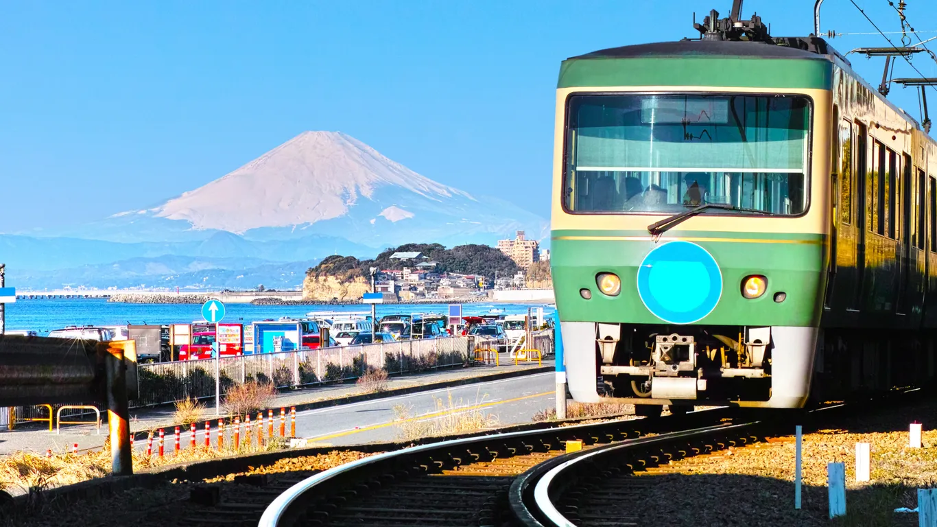 Ein grün-cremefarbener Zug der Enoden-Bahn fährt am Meer entlang, im Hintergrund der Fuji mit Schneekappe